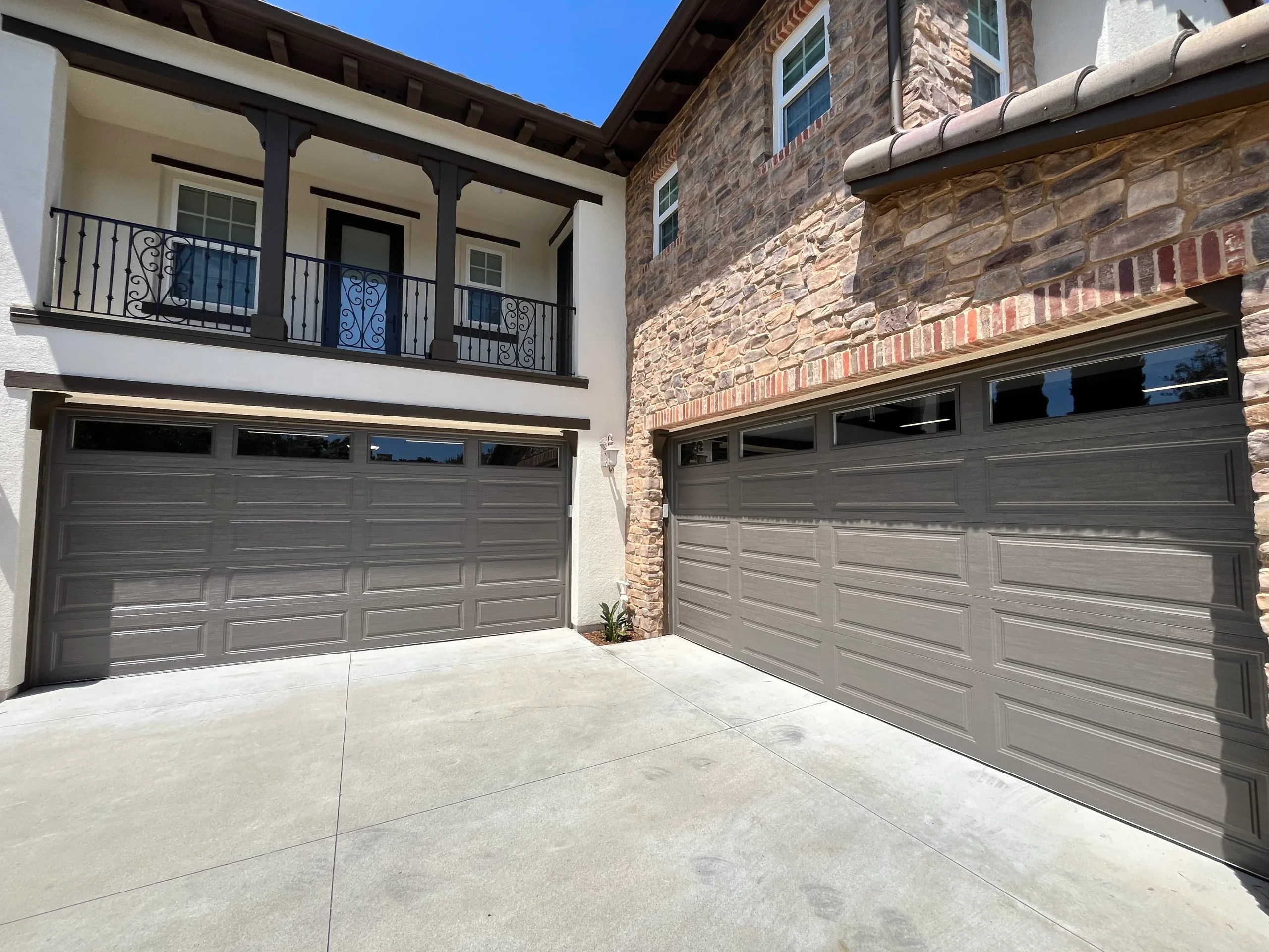 An exterior view of a multi-story stone and stucco home with two large, tan sectional doors, ideal for referencing estimates on garage door repair cost.