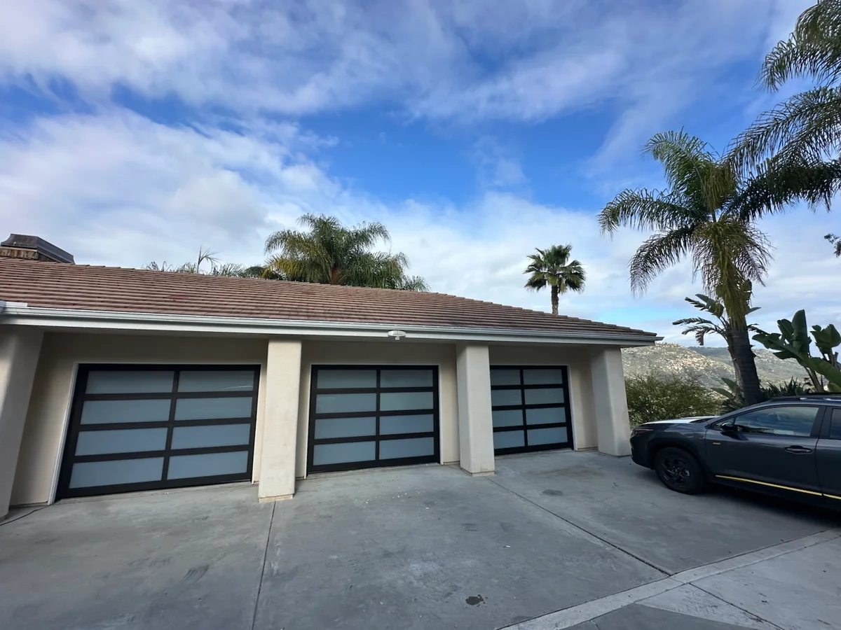 An exterior view of a three-car garage featuring modern full-view frosted glass doors with black aluminum frames, perfect for answering how much does garage door repair cost.