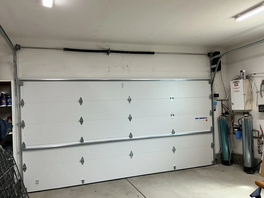 An interior view of a white garage door showing the high-tension torsion spring and tracks, emphasizing crucial components to inspect for hidden garage door hazards.