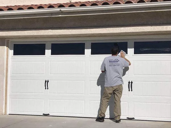 A technician cleaning the window panels of a white residential garage door on a sunny day, demonstrating routine garage door maintenance during summer.