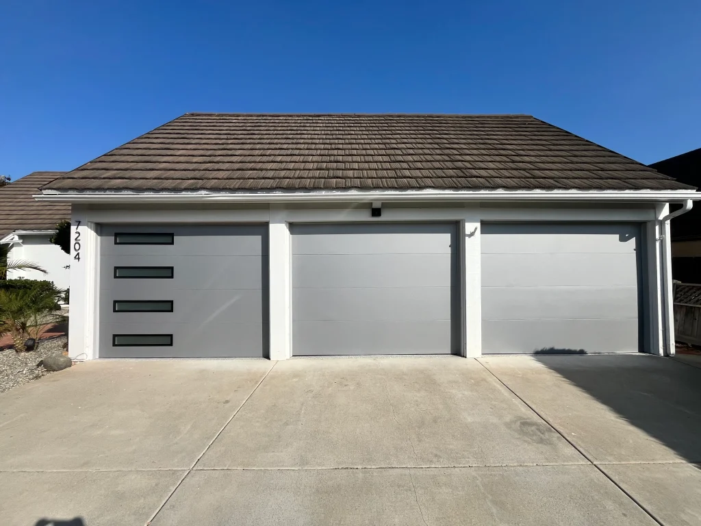 An exterior view of a three-car garage with modern gray flush-panel doors under a clear blue sky, highlighting a perfect time for a summer garage door service schedule.