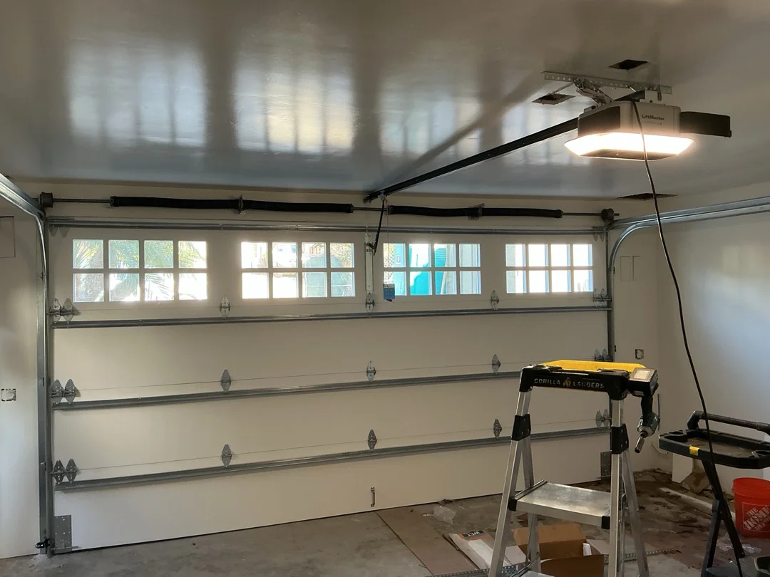 An indoor view of a garage featuring a white door with square window panels, torsion springs, and an overhead LiftMaster opener, set up for a professional summer garage door service.