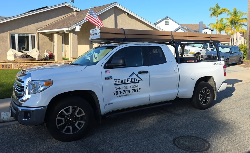 A white Bradbury Garage Doors company truck parked on a residential street with equipment loaded on its roof rack, representing professional garage door maintenance near me.