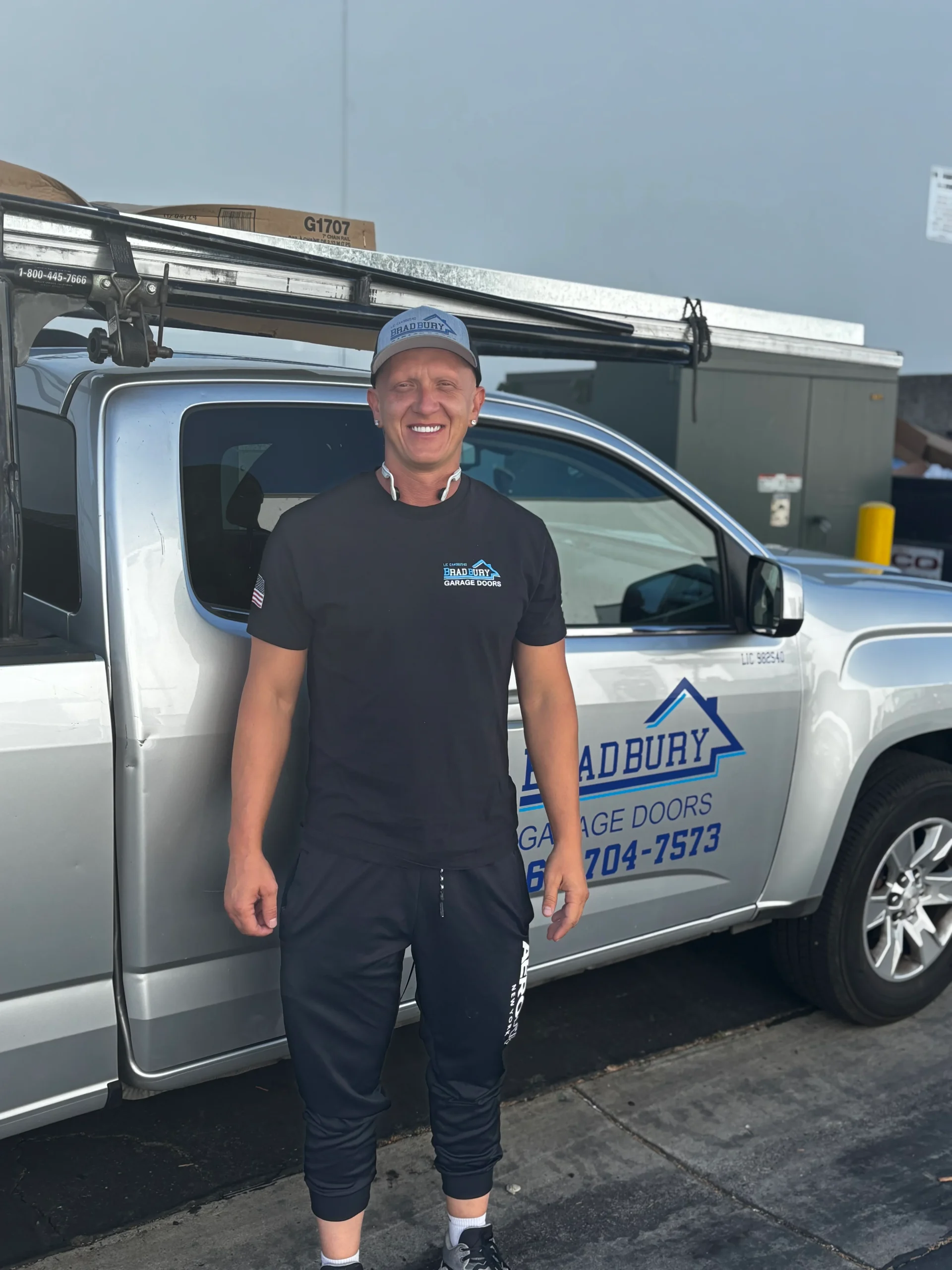 A smiling Bradbury Garage Doors technician standing in front of a silver company work truck, ready to perform a professional garage door tune up.